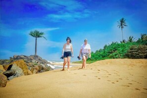 Dos personas caminando sobre una playa arenosa, con palmeras y rocas al fondo.