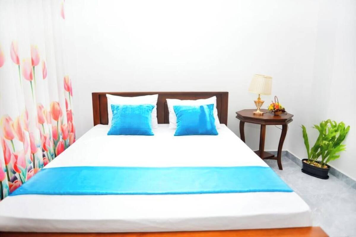 Una cama con cabecera de madera, dos almohadas, una mesita de noche con una lámpara y una pequeña planta en maceta.