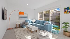 Un moderno salón con un sofá azul, una mesa de centro blanca y un amplio ventanal con persianas.
