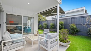 Un patio con mobiliario blanco, una puerta de cristal que da a una habitación y una valla gris al fondo.