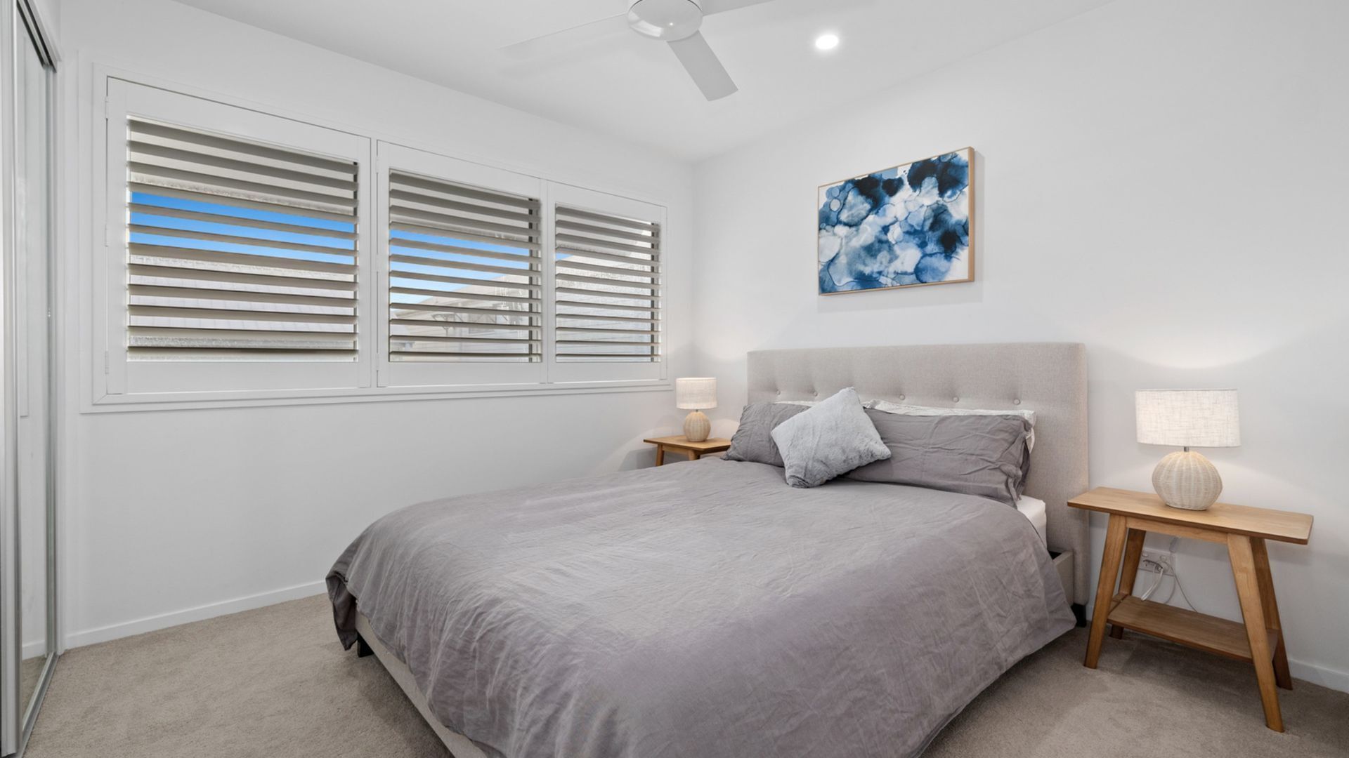 Un dormitorio con una cama, dos mesitas de noche con lámparas, un ventilador de techo y una ventana con persianas.