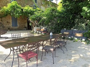 Une terrasse extérieure avec une longue table et des chaises, entourée de verdure et bordée d’un bâtiment en pierre.