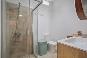 A bathroom with a glass shower enclosure, a white toilet, a green trash bin, and a wooden vanity with a sink.