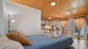 A bedroom with a bed, pillows, a bedside lamp, a wooden ceiling, a window with curtains, and a view of the outdoors.