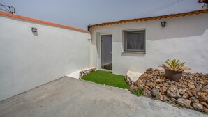 A small, white-walled building with a tiled roof, a door, a window, and a potted plant.