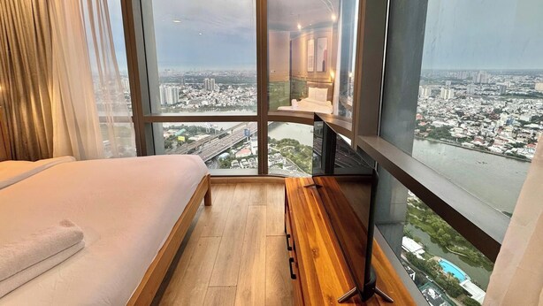 A hotel room with a large window offering a city view, a bed, a wooden bench, and a bedside table. - The Landmark 81 Deluxe Apartment Suite (Ho Chi Minh City)