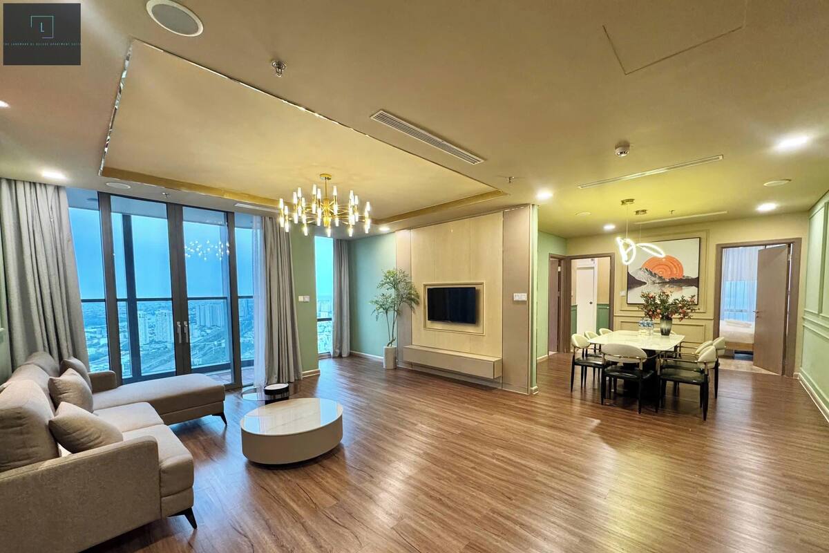 Un salón amplio con un sofá grande, una mesa de centro redonda y un comedor con mesa y sillas. Un televisor de pantalla plana está montado en la pared, y hay un ventanal con vistas a la ciudad.