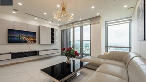 A modern living room with a large flat-screen TV, a black coffee table with a vase of flowers, a beige sofa, and a large window offering a city view. - The Landmark 81 Deluxe Apartment Suite (Ho Chi Minh City)