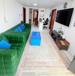 Une pièce avec un canapé vert, un pouf bleu, un téléviseur et un bureau avec une chaise.
