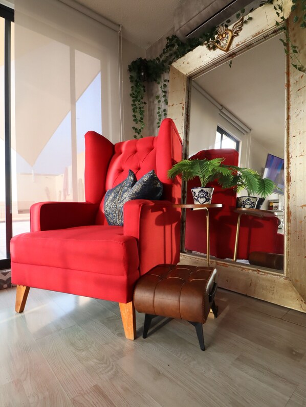 Un fauteuil rouge à haut dossier et un pouf marron, une petite table sur laquelle est posée une plante en pot, et un grand miroir qui reflète la pièce.