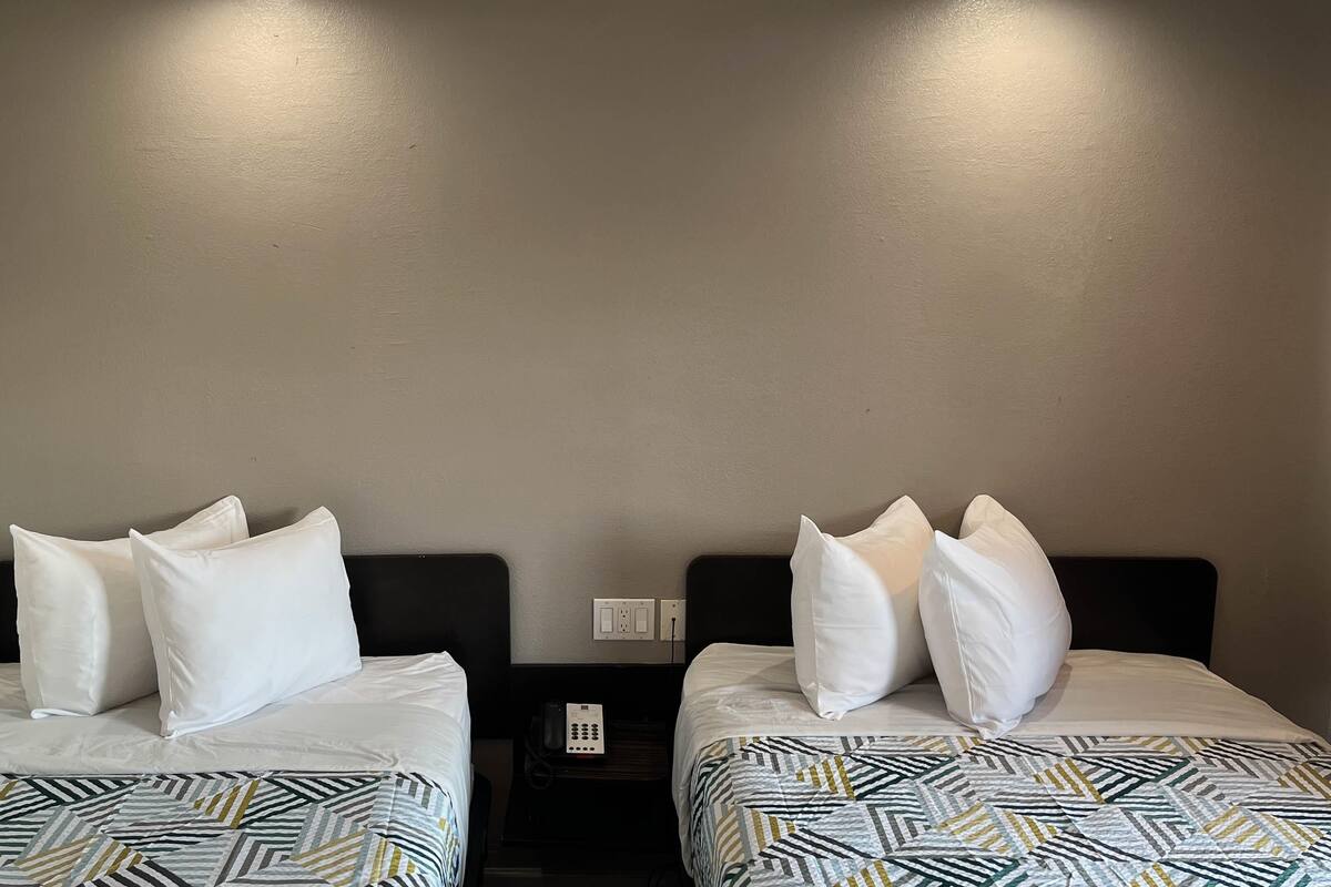 A hotel room with two beds, each with white bedding and patterned quilts. There is a bedside table with a phone between the beds.