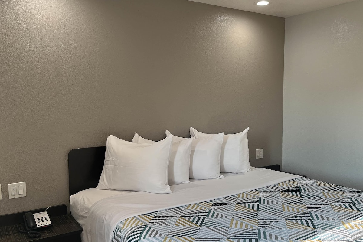 A hotel room with a bed, white pillows, a patterned bedspread, a telephone on the nightstand, and a wall socket.