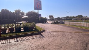A motel sign with the number 6, a paved entrance, and a fenced area with greenery. - Motel 6 Dallas, Tx – St. Augustine / Hwy 175 (Dallas)