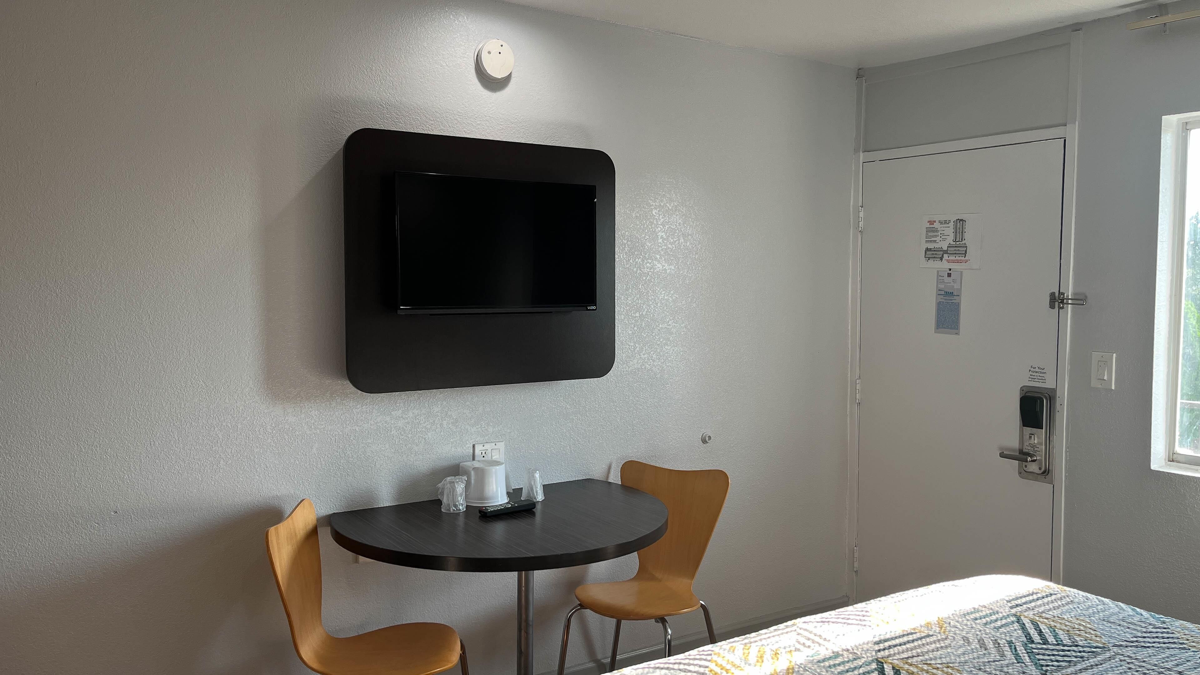 Een hotelkamer met een flatscreen televisie aan de muur, een klein rond tafeltje met twee stoelen, en een deur met een raam.