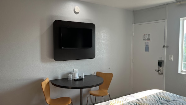 A hotel room with a flat-screen TV mounted on the wall, a small round table with two chairs, and a door with a window.
