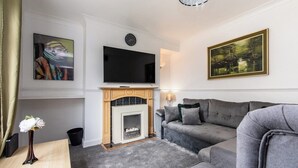 A living room with a fireplace, a grey sofa, a flat-screen TV mounted above the fireplace, and a painting on the wall.