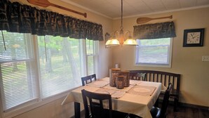 Comedor con mesa dispuesta para cuatro, sillas de madera, una lámpara de araña y dos ventanas con persianas.