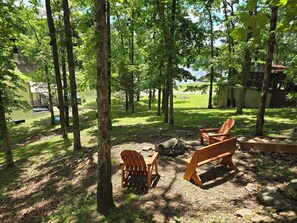 A serene outdoor area with wooden chairs and a small wooden structure by a lake, surrounded by trees.