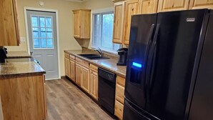 Una cocina con armarios de madera, un refrigerador negro, lavavajillas y una ventana con persianas.