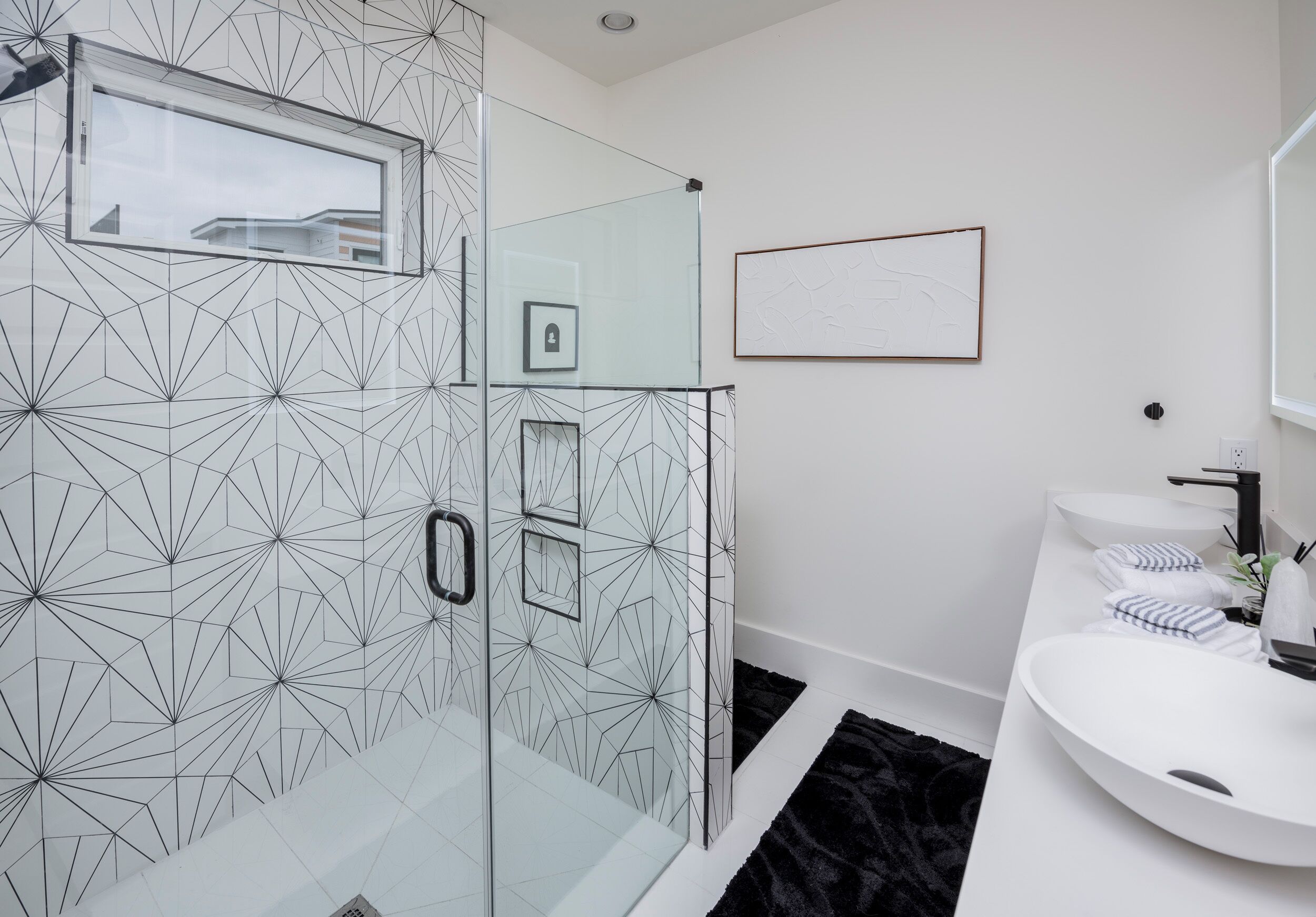 Un baño moderno con pared de diseño geométrico, cabina de ducha con mampara de vidrio, lavamanos blanco y un cuadro en la pared.