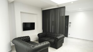A modern living room with two black leather sofas, a flat-screen TV mounted on the wall, and a dark wooden panel wall.