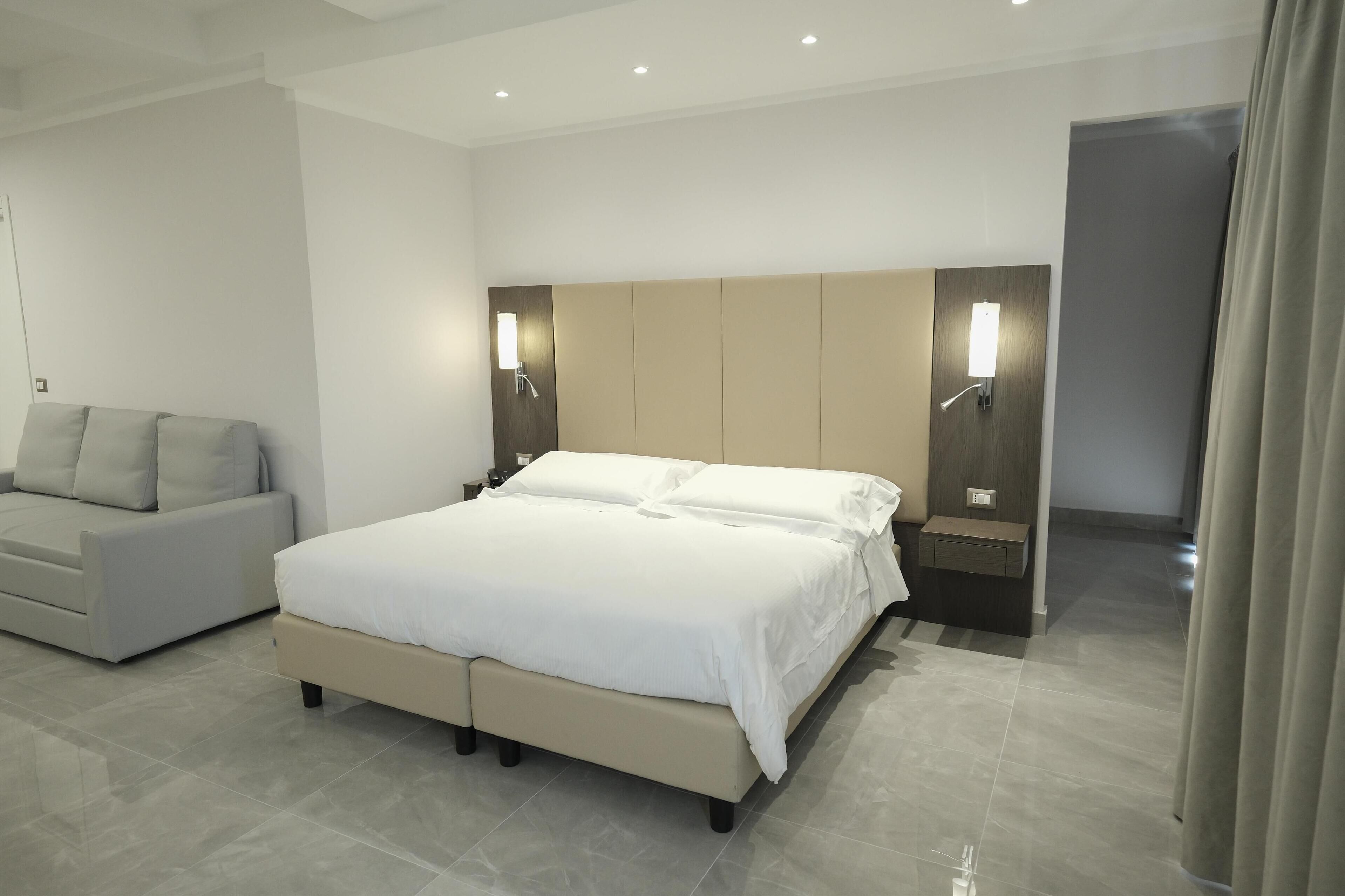 A modern hotel room with a large bed, a sofa, and two wall-mounted lights.