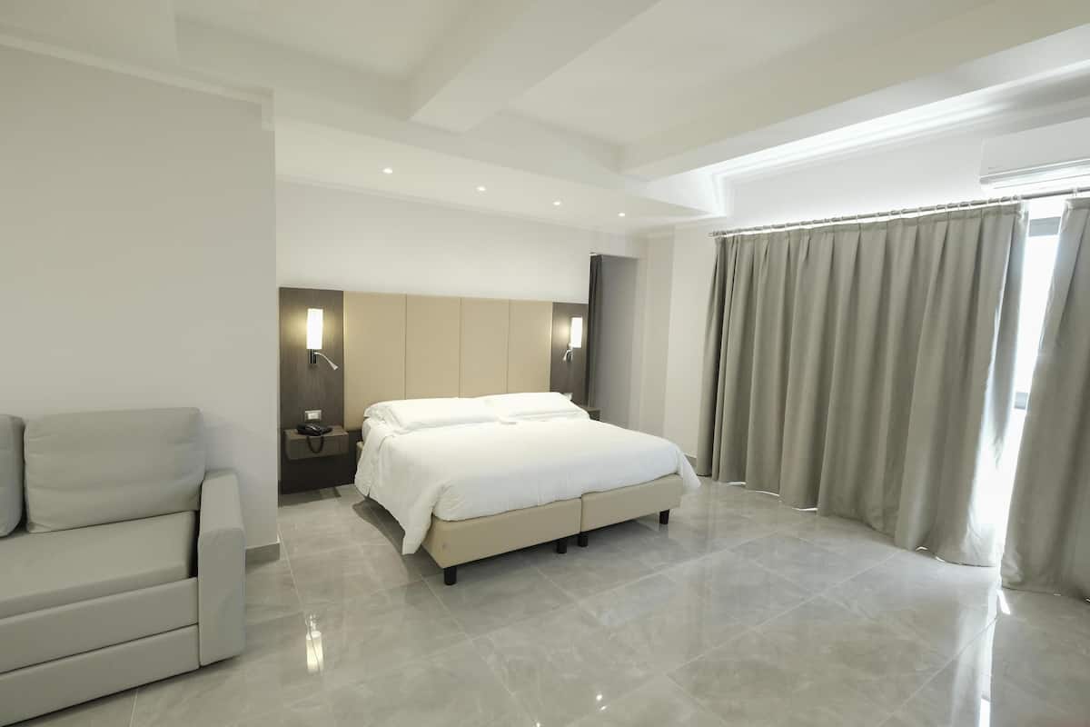 A modern hotel room with a large bed, a sofa, a bedside table, and a mirror.