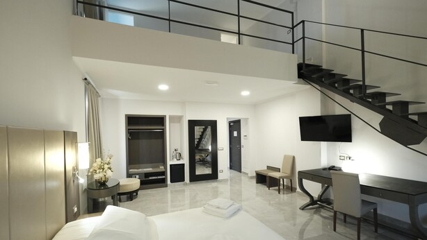 A modern hotel room with a large bed, a desk, a chair, a TV, a staircase, and a bathroom.