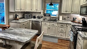 A kitchen with white cabinets, a marble countertop, a dishwasher, a stove, and a window with a view of trees. - Lakefront Retreat + Dock + Kayaks + Fire Pit + Lakefront Kitchen (Milledgeville)