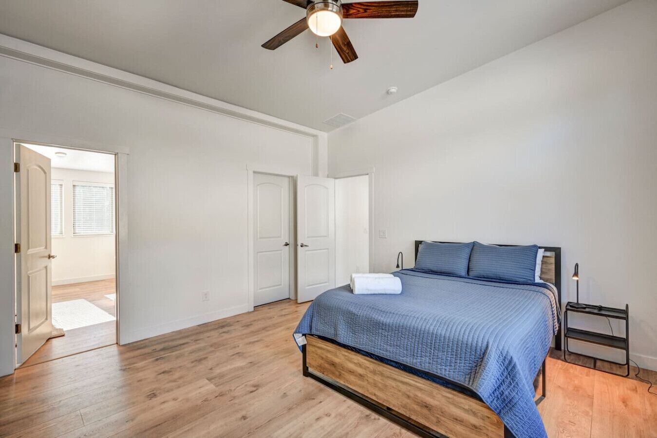 A bedroom with a wooden floor, a bed with a blue bedspread, a ceiling fan, and a nightstand with a lamp.