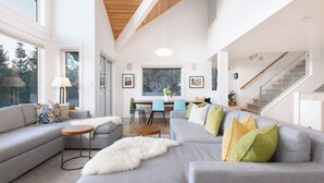 Een moderne woonkamer met een grijze bank, een ronde houten salontafel en een witte bontmantel. Er is een zithoek met een tafel en stoelen, en een trap die naar een ander niveau leidt.