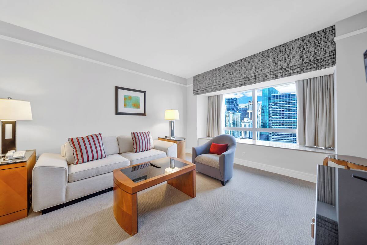A modern hotel room with a sofa, armchair, and a coffee table. There is a window with a city view, a lamp, and a framed picture on the wall.