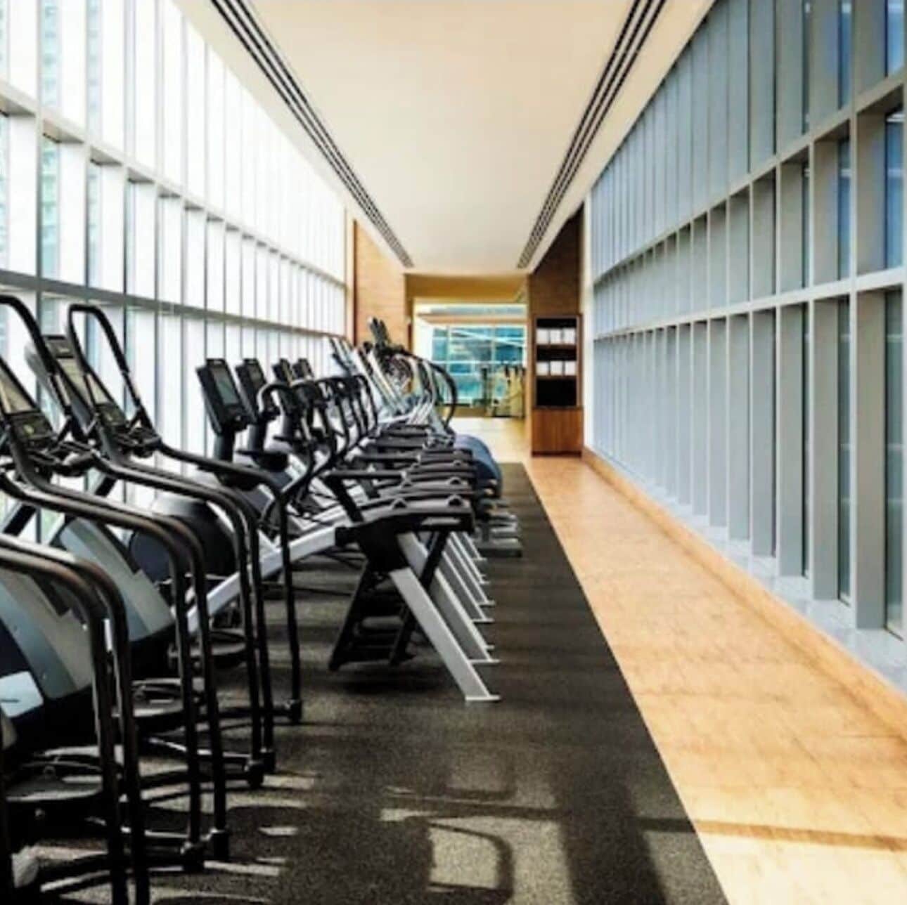 Un gimnasio con cintas de correr alineadas a lo largo de un pasillo, con grandes ventanales en un lateral.