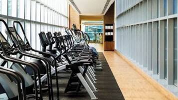 Un gimnasio con cintas de correr alineadas a lo largo de un pasillo, con grandes ventanales en un lateral.