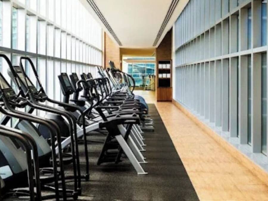 Un gimnasio con cintas de correr alineadas a lo largo de un pasillo, con grandes ventanales en un lateral.