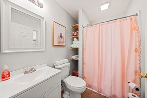 Une salle de bain avec un lavabo blanc, des toilettes et un rideau de douche. Un tableau encadré est accroché au mur, et des étagères contiennent des serviettes et des articles de toilette.