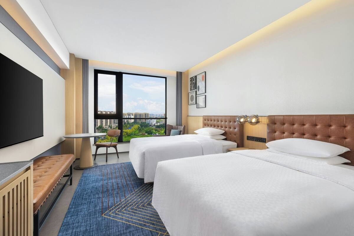A hotel room with two beds, a TV, a window with a city view, and a desk with a chair.