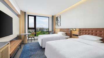 A hotel room with two beds, a TV, a window with a city view, and a desk with a chair.