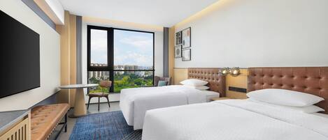 A hotel room with two beds, a TV, a window with a city view, and a desk with a chair.