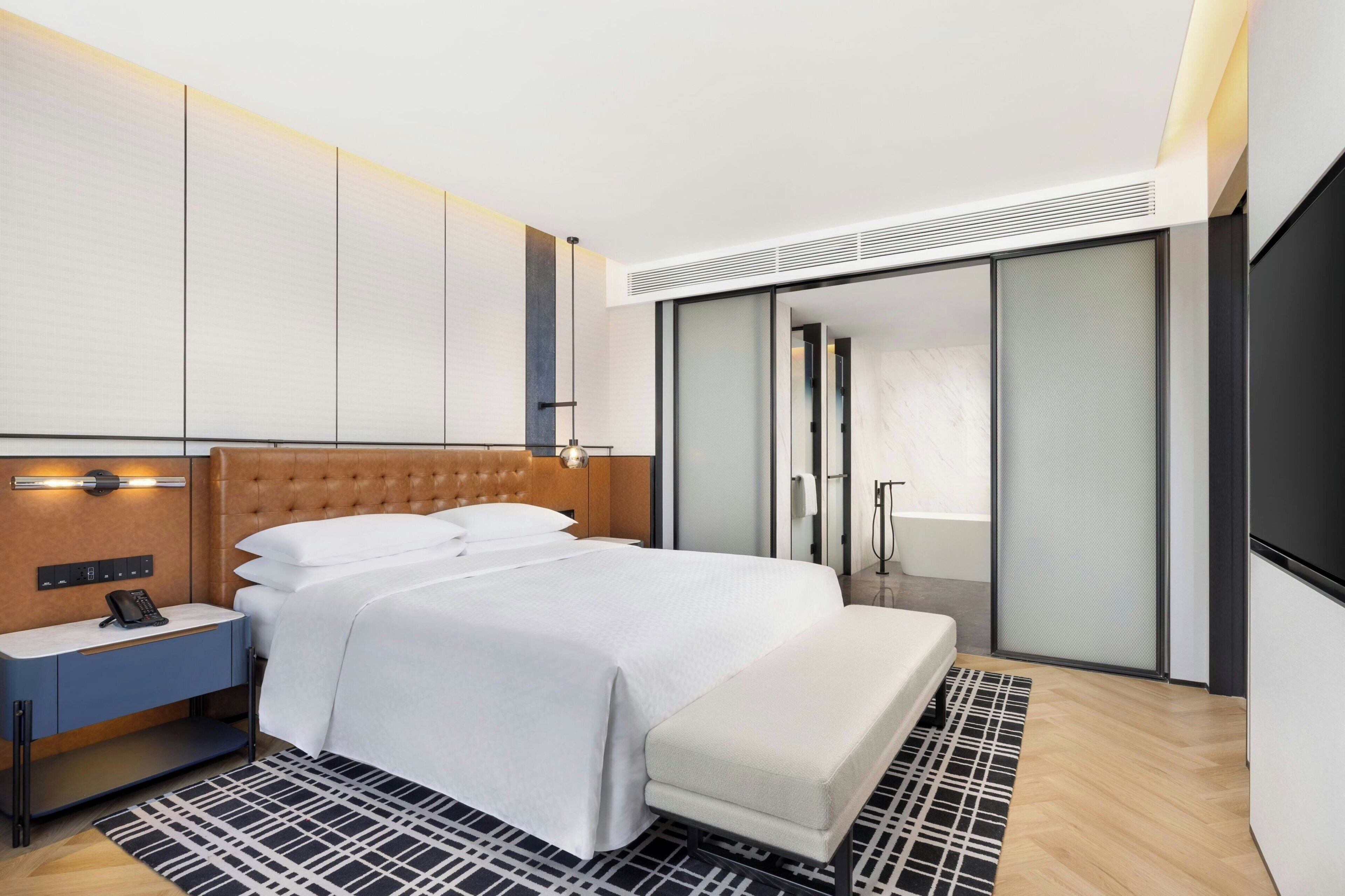 A modern hotel room with a large bed, a bedside table with a phone, a bench, a patterned rug, and a sliding door closet.