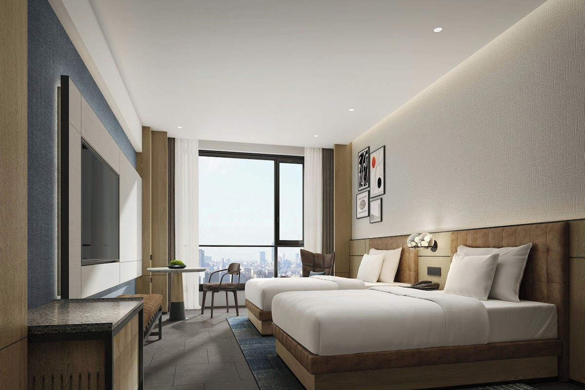 A modern hotel room with two beds, a desk, a chair, and a large window offering a city view.