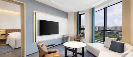Habitación de hotel moderna con un televisor grande, un sofá, una silla, una mesita y vistas a la ciudad.