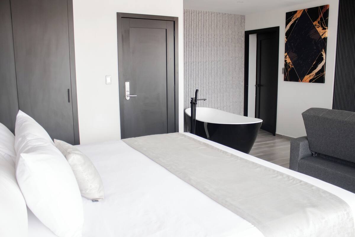 Une chambre d’hôtel moderne avec un grand lit, un canapé, une baignoire noire et une grande fenêtre.