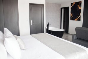 Una habitación de hotel moderna con una cama grande, un sofá, una bañera negra y un ventanal amplio.