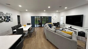 A modern living room with a dining area, a large TV, and a sliding glass door leading to a garden. - Enjoy our brand new home close to Hollywood in Hallandale Beach! (Hallandale Beach)