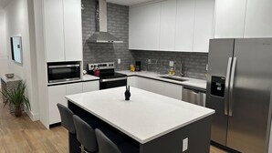 A modern kitchen with white cabinets, stainless steel appliances, a central island with bar stools, and a grey tiled backsplash. - Enjoy our brand new home close to Hollywood in Hallandale Beach! (Hallandale Beach)
