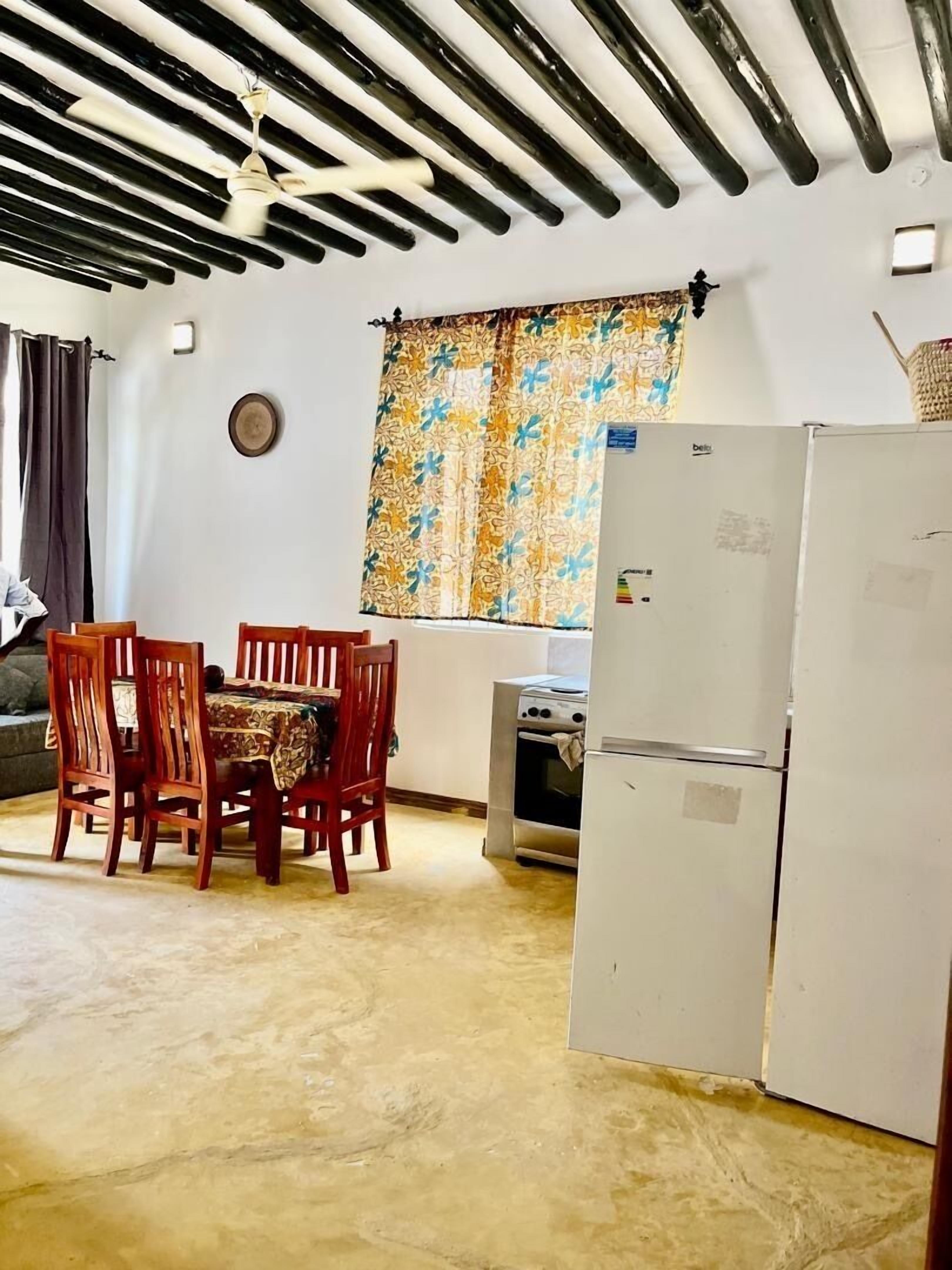 A compact kitchen with a dining table and chairs, a refrigerator, and a window with patterned curtains.