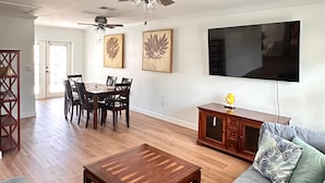 Un salon spacieux doté d’un parquet, d’un canapé gris, d’une table basse en bois, d’un coin repas avec une table et des chaises en bois, et d’un ventilateur de plafond.