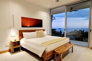 A bedroom with a large bed, a bedside table, a bench, a painting, and a view of the sea. - Casa Playitas: Luxury 5BR Villa in Manuel Antonio (Quepos)