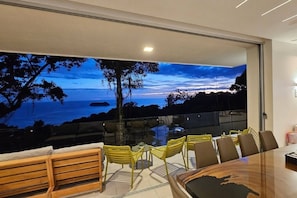 A modern dining area with a panoramic ocean view, a glass partition, and a wooden table with chairs. - Casa Playitas: Luxury 5BR Villa in Manuel Antonio (Quepos)